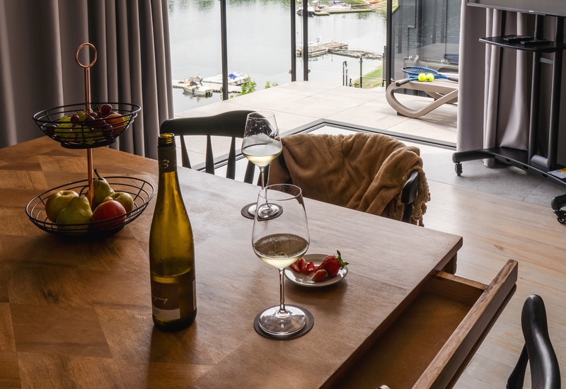 Esstisch mit Wein, Gläsern und Obst, kuschelige Decke auf dem Stuhl, offener Balkon mit Blick auf Marina und grüne Hügel in moderner Suite am Wasser