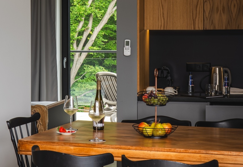 Holztisch mit Weingläsern und Obstkorb, schwarze Stühle, Küchenzeile mit Wasserkocher und Kaffeemaschine, Balkonblick auf grüne Bäume