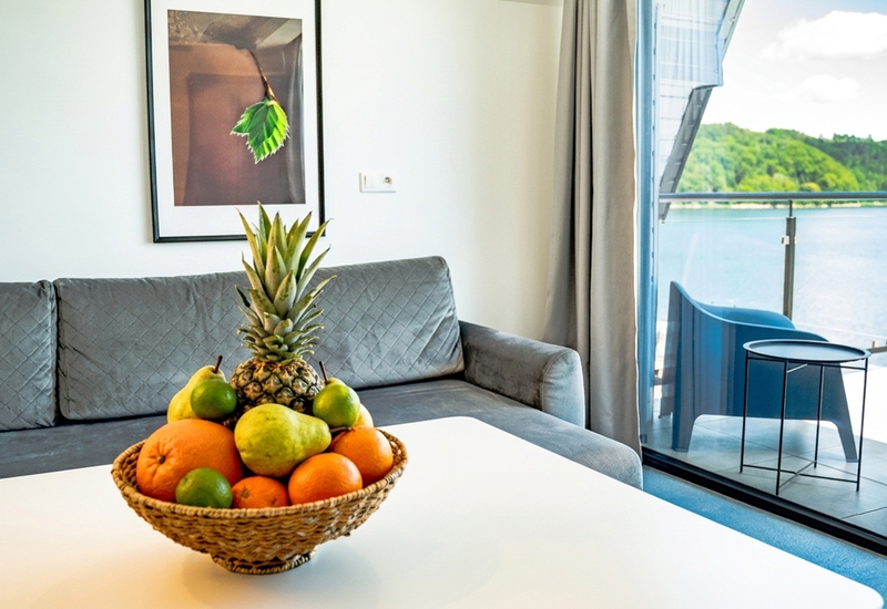 Heller Wohnbereich mit grauem Sofa, Obstkorb auf dem Tisch, Balkon mit See- und Hügelblick