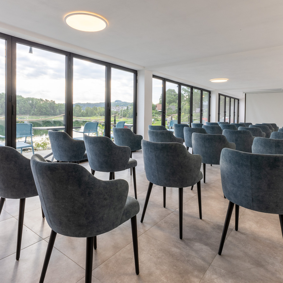 Konferenzraum mit gepolsterten Sesseln, Leinwand und bodentiefen Fenstern mit Blick auf die Seeterrasse.