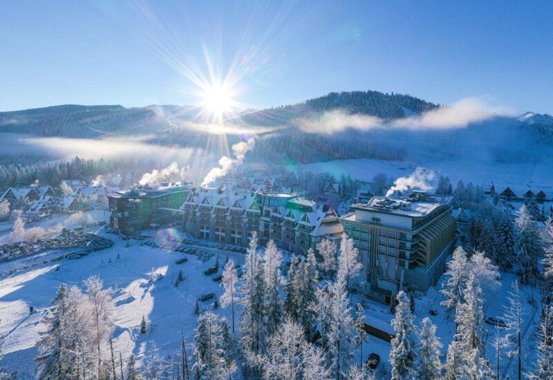 Zasnežený hotel v horách videný z vtáčej perspektívy, s panorámou Tatier a slnečnými lúčmi nad vrcho