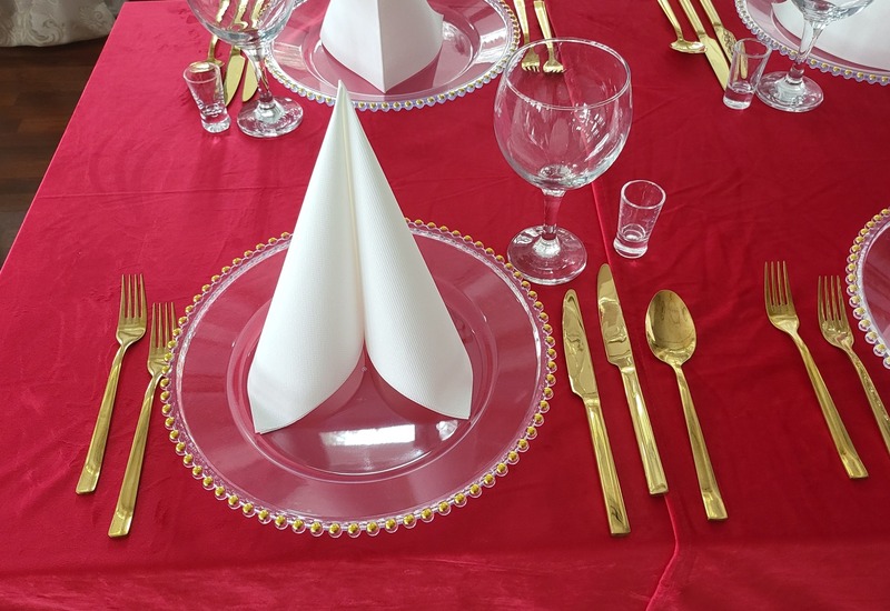 Eleganckie nakrycie: czerwony obrus, złote sztućce, przezroczyste talerze z koralikową krawędzią, stojące białe serwetki.