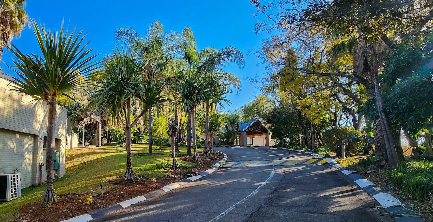 Palm-lined hotel driveway leading to a covered reception pavilion, manicured lawn, tropical trees and sunlit blue sky.