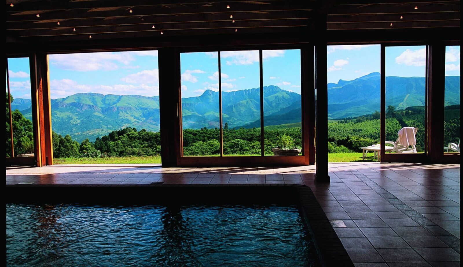 Indoor pool with tiled deck and wooden beams, floor-to-ceiling glass opening to lawn, lounge chairs and mountain vistas.