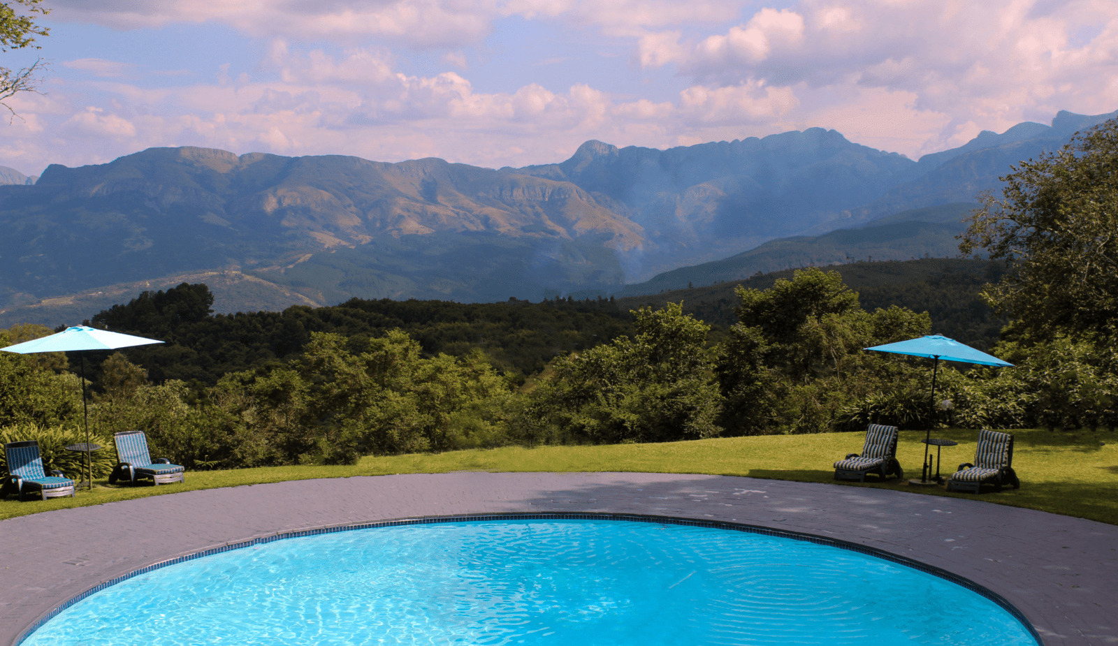 Round outdoor pool with clear blue water, striped lounge chairs and umbrellas on a lawn, panoramic mountain vista and forested valley beyond