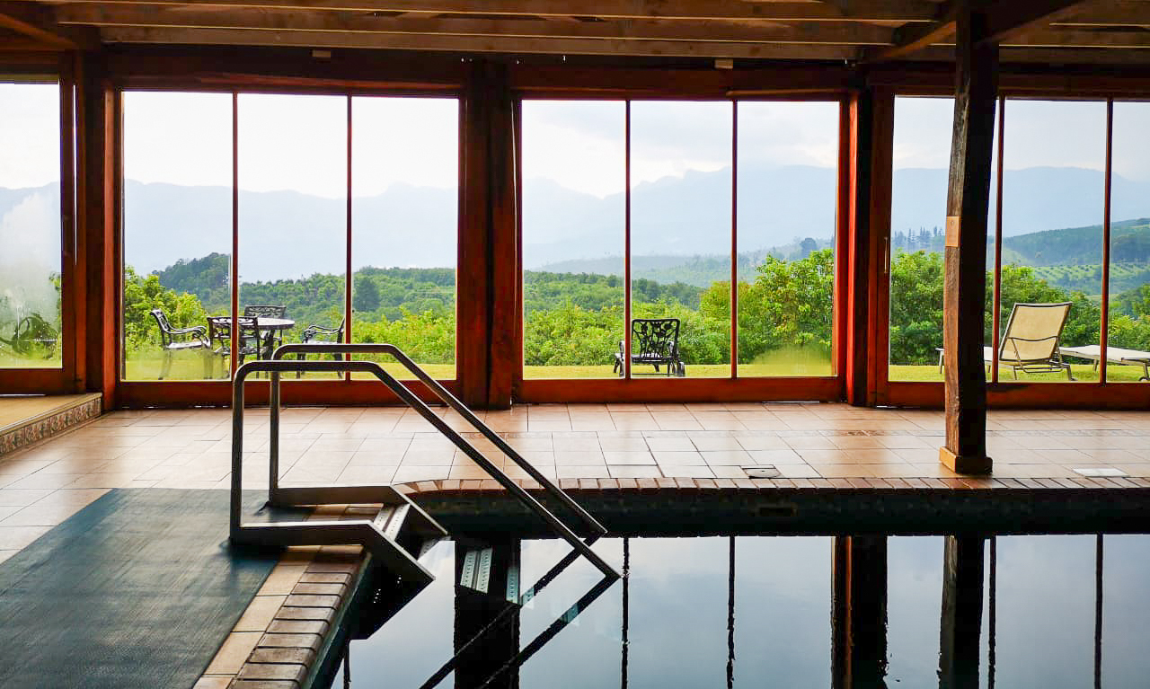 Indoor pool with handrail and tiled deck, floor-to-ceiling windows framing mountain vista and patio seating