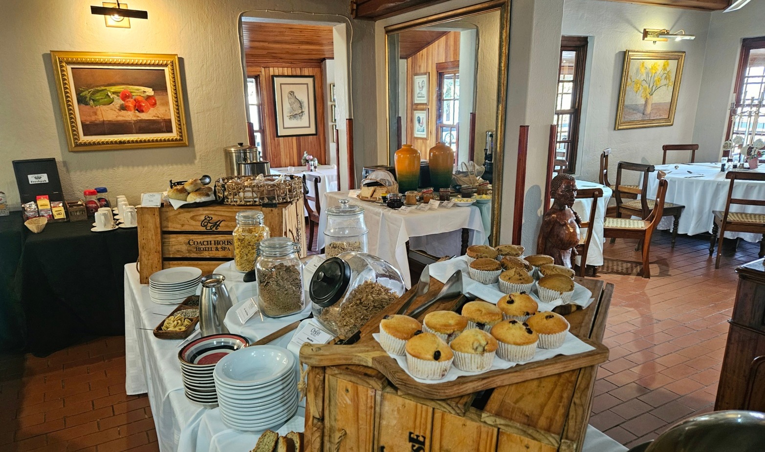 Hotel breakfast buffet with muffins, cereals in glass jars, plates and coffee on rustic crates in a cozy wood-paneled dining room.