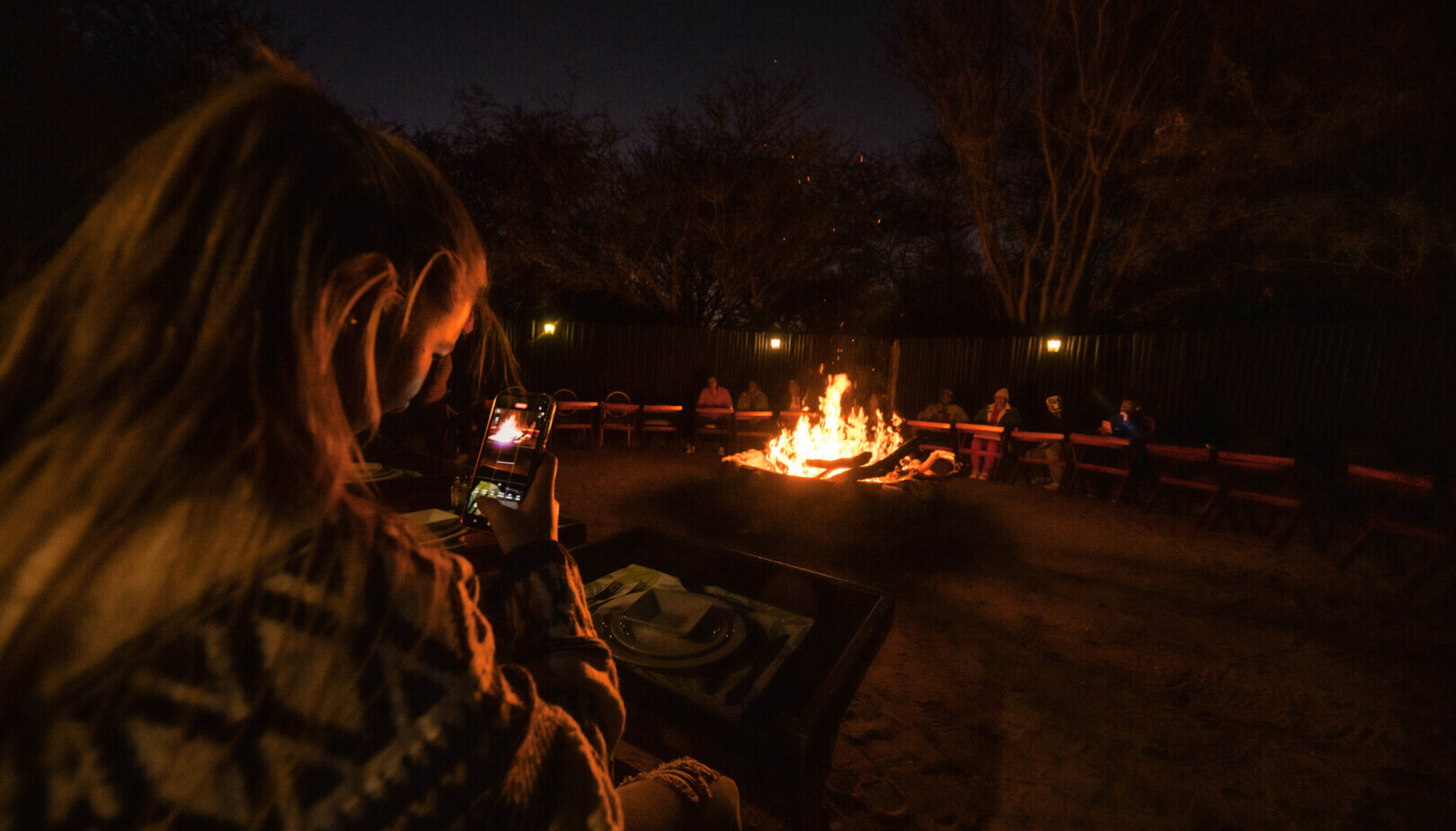 Guest photographing a night fire pit with communal benches and table settings in a lodge courtyard, warm, social atmosphere.