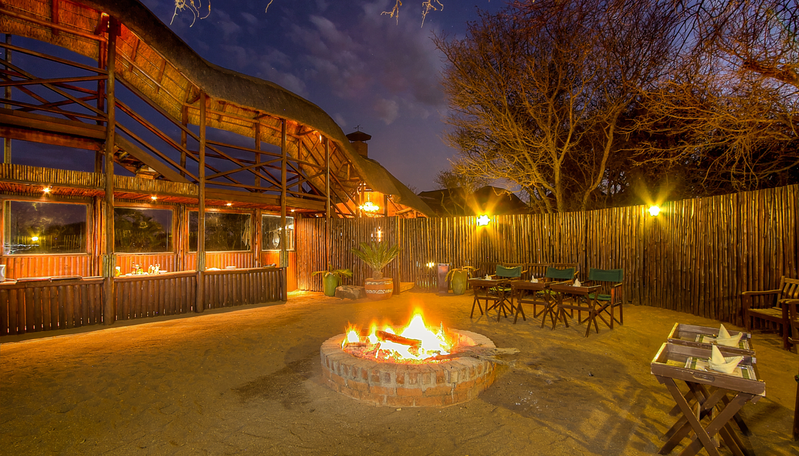 Thatched-roof lodge courtyard at night with a brick campfire, wooden dining tables and warm ambient lighting.