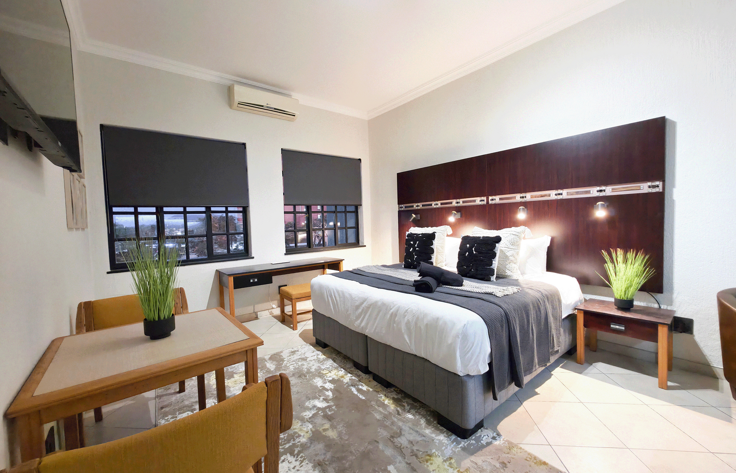 Modern double hotel room with king bed, dark wood headboard, bedside lamps, desk, dining table, city-view windows and AC.