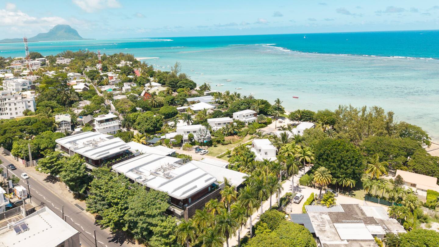 Aerial view of palm-lined beachfront with modern white villas and resort buildings facing turquoise lagoon and anchored boats.