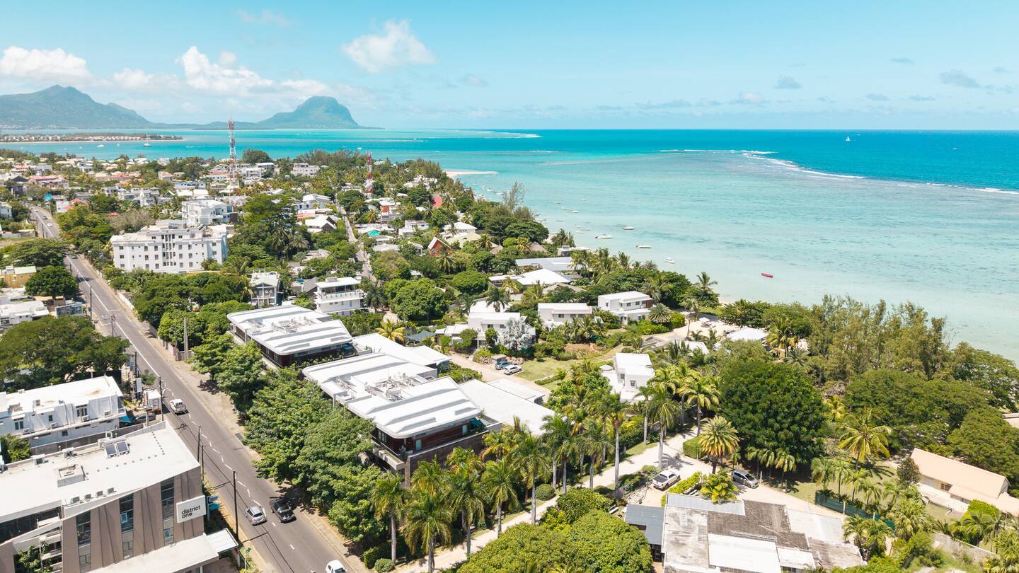Coastal resort town with palm-lined avenue, low-rise white villas and hotels beside a turquoise lagoon and distant mountains.