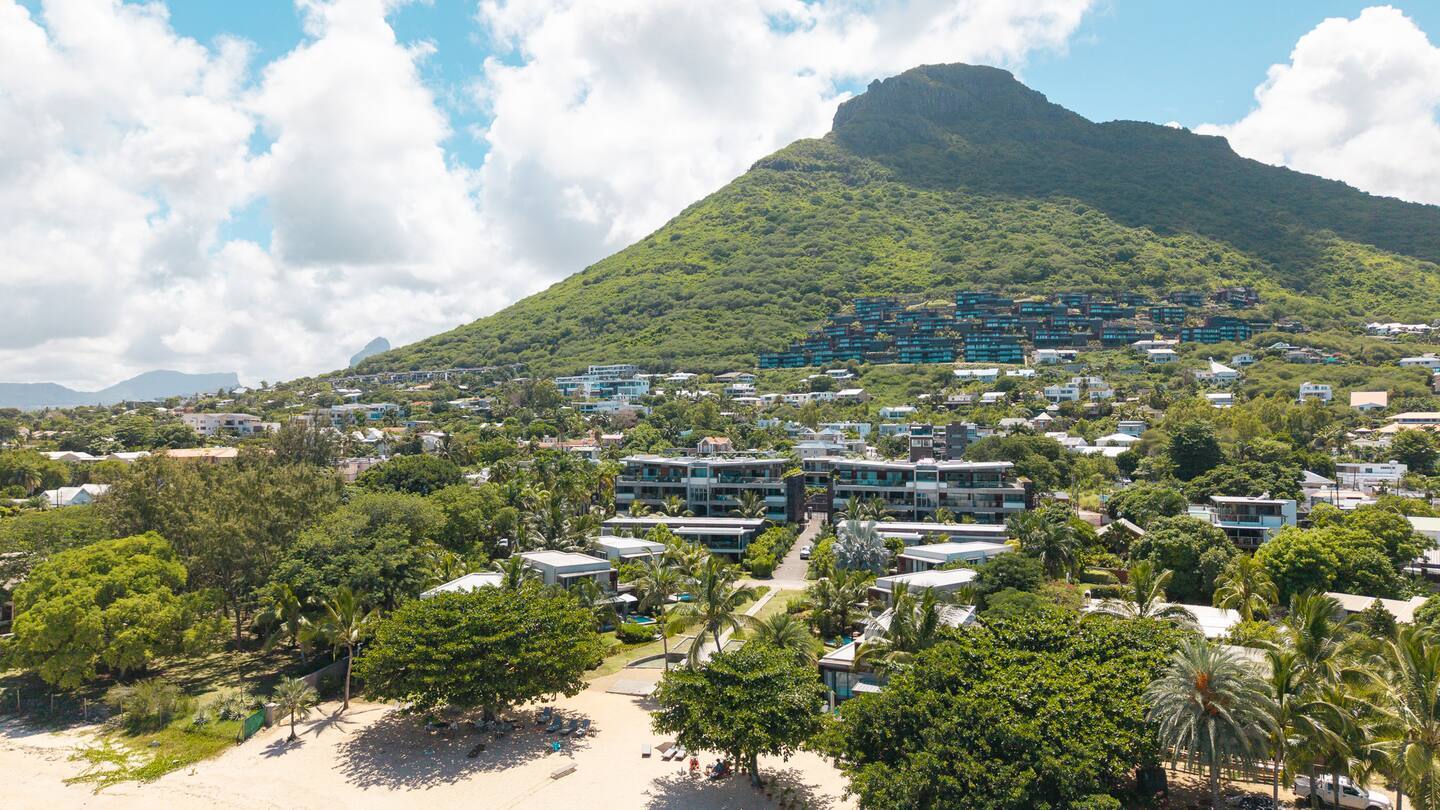 Beachfront resort with modern low-rise villas and palm-lined sandy beach, set below a lush green mountain