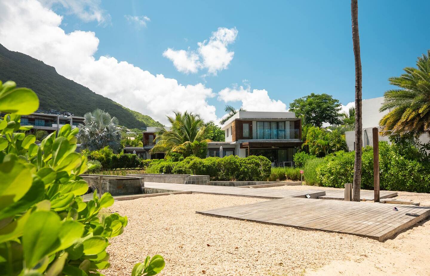 Modern beachfront villa with private wooden deck, palm trees and lush gardens, mountain backdrop under bright blue sky