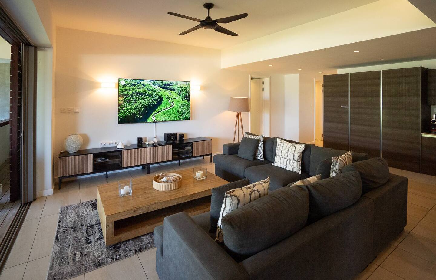 Spacious modern suite living area with large dark sectional, wooden coffee table, wall-mounted TV, media console, floor lamp and balcony.