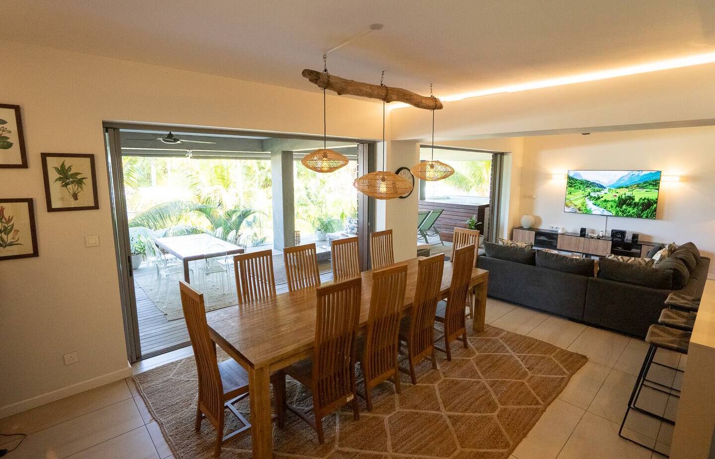 Open-plan dining and lounge with 10-seat wooden table, rattan pendant lights, large sofa and sliding doors to a palm-lined terrace