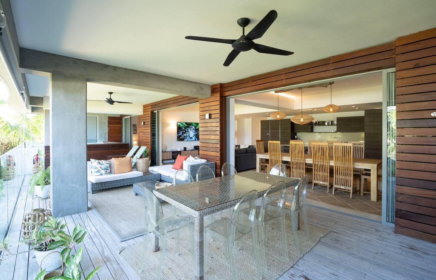 Covered tropical terrace with wicker dining table and clear chairs, lounge sofas, ceiling fans, and sliding doors to indoor dining and kitchen.