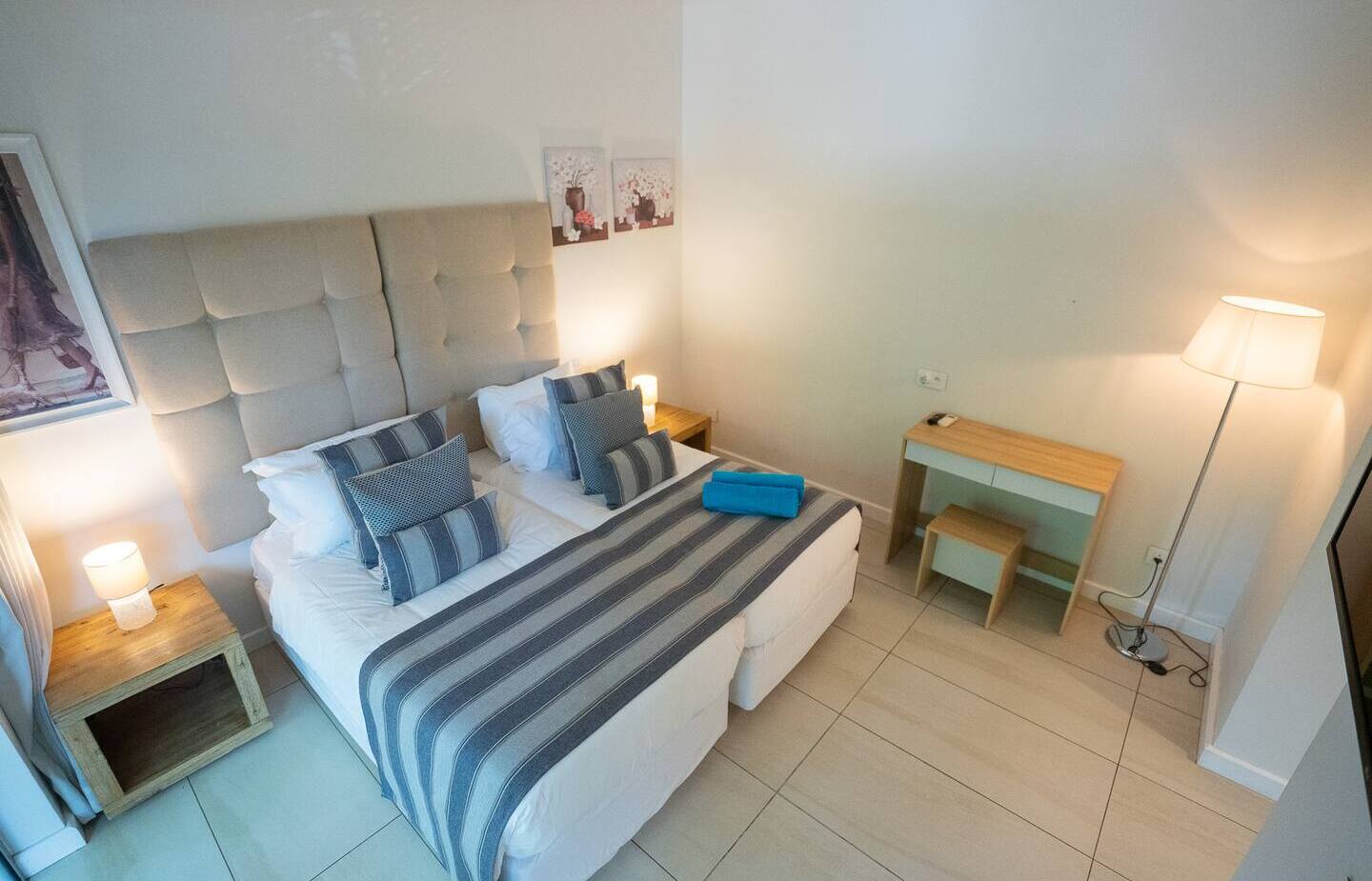 Bright modern twin-bed room with tufted headboard, striped bed runner and pillows, bedside lamps, wooden desk, floor lamp and folded turquoise towels.