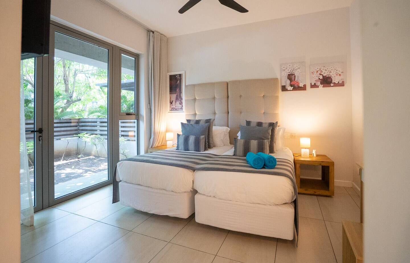Bright modern twin-bed room with tufted headboard, striped linens, turquoise rolled towels, bedside lamps, ceiling fan and patio doors to garden.