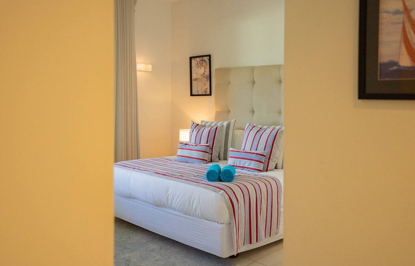 Bright guest bedroom with tufted headboard, striped red-and-blue bedding, bedside lamp and two rolled turquoise towels.