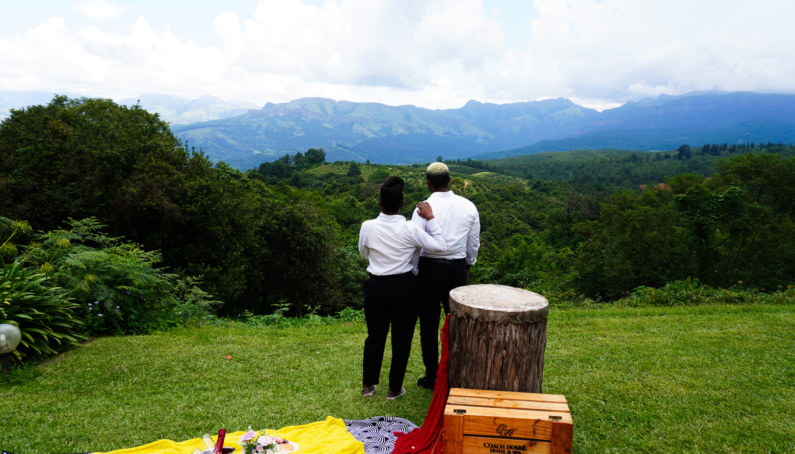 Two people in white shirts embrace while overlooking a lush green valley and blue mountains in Tzaneen, Limpopo, with a picnic setup, wine and a Coach House Hotel & Spa wooden crate in the foreground.