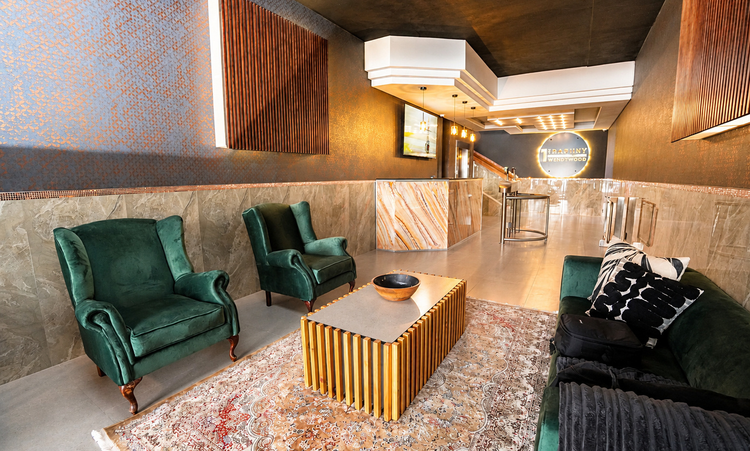 Boutique lobby with marble reception desk, warm pendant lights, green velvet seating, wooden slat accents, patterned rug and sleek coffee table.
