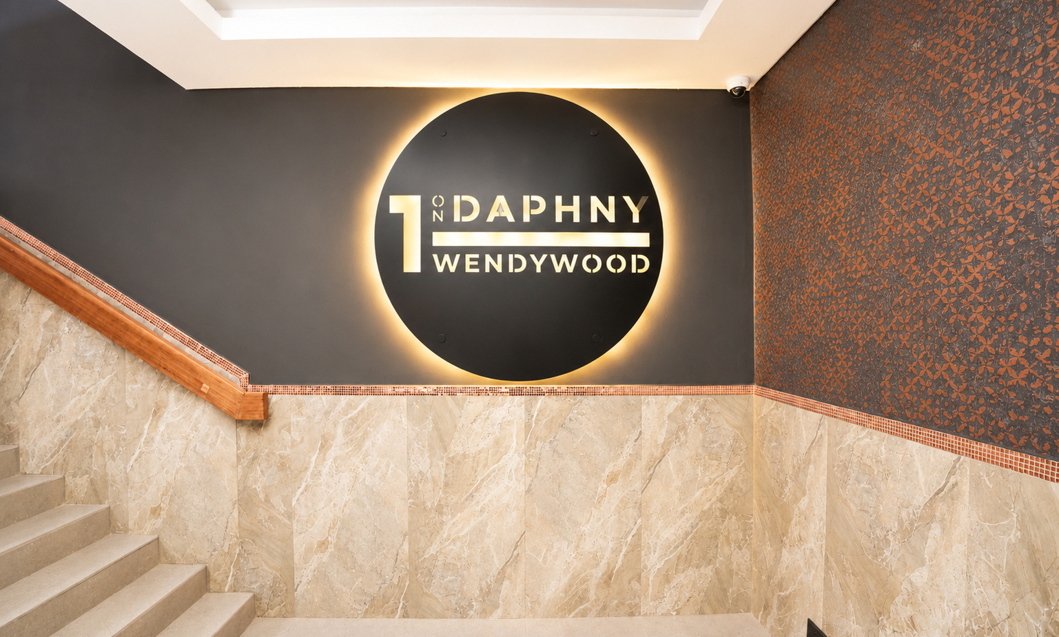 Backlit circular lobby sign reading 1 ON DAPHNY WENDYWOOD above marble stairs with wood handrail and patterned accent wall, warm light