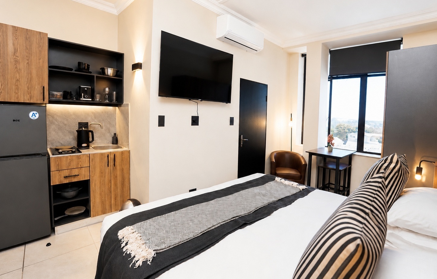 Studio hotel room: queen bed, striped bolster, wall TV, kitchenette with mini-fridge and coffee maker, city-view window.