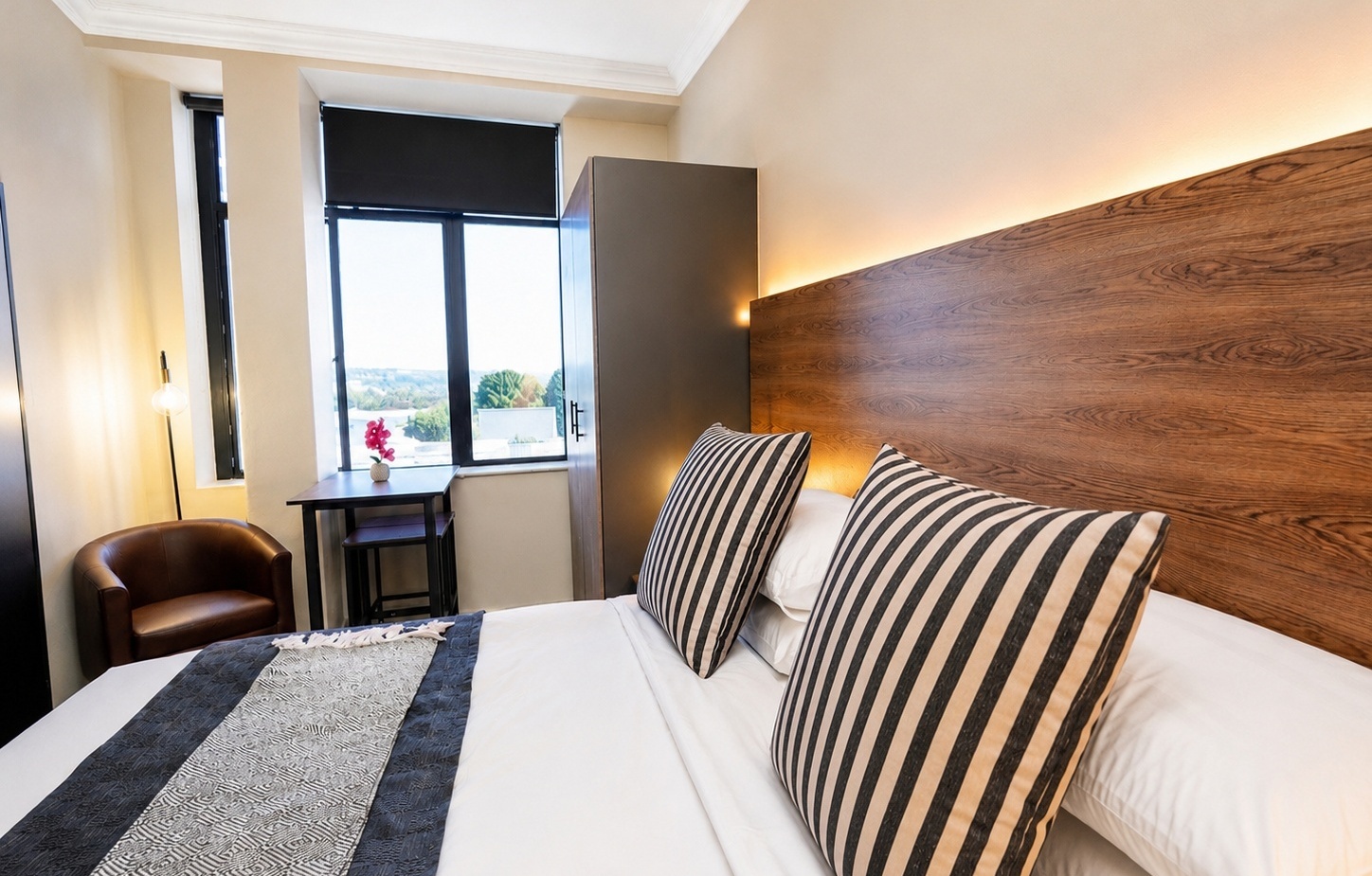 Contemporary hotel room with wooden headboard with backlighting, striped pillows on white bed, window view, desk with orchid.