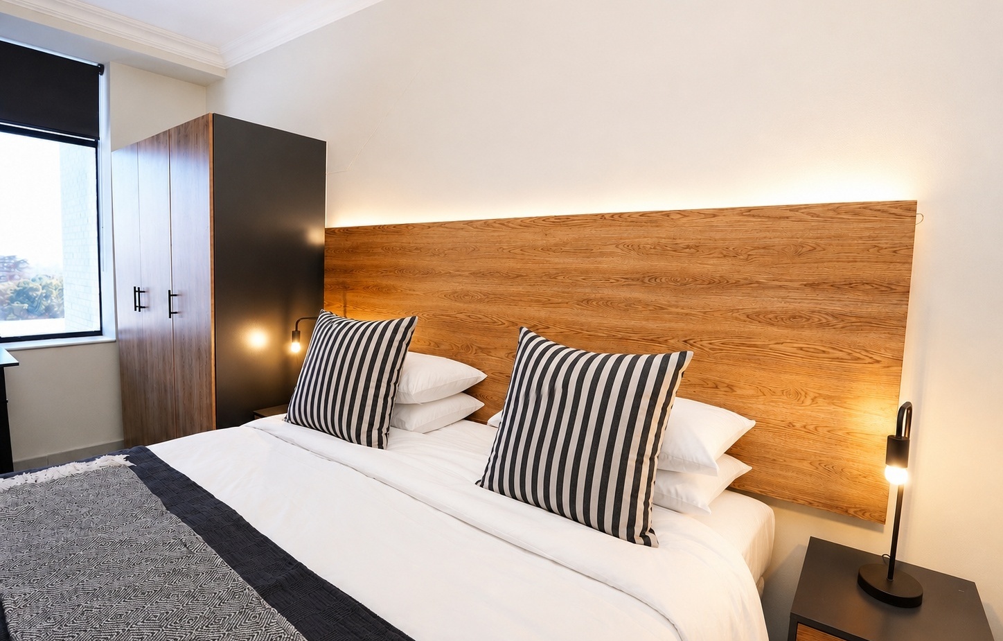 Modern hotel king bed with wood headboard, black-and-white striped pillows, bedside lamps, wardrobe and natural light.