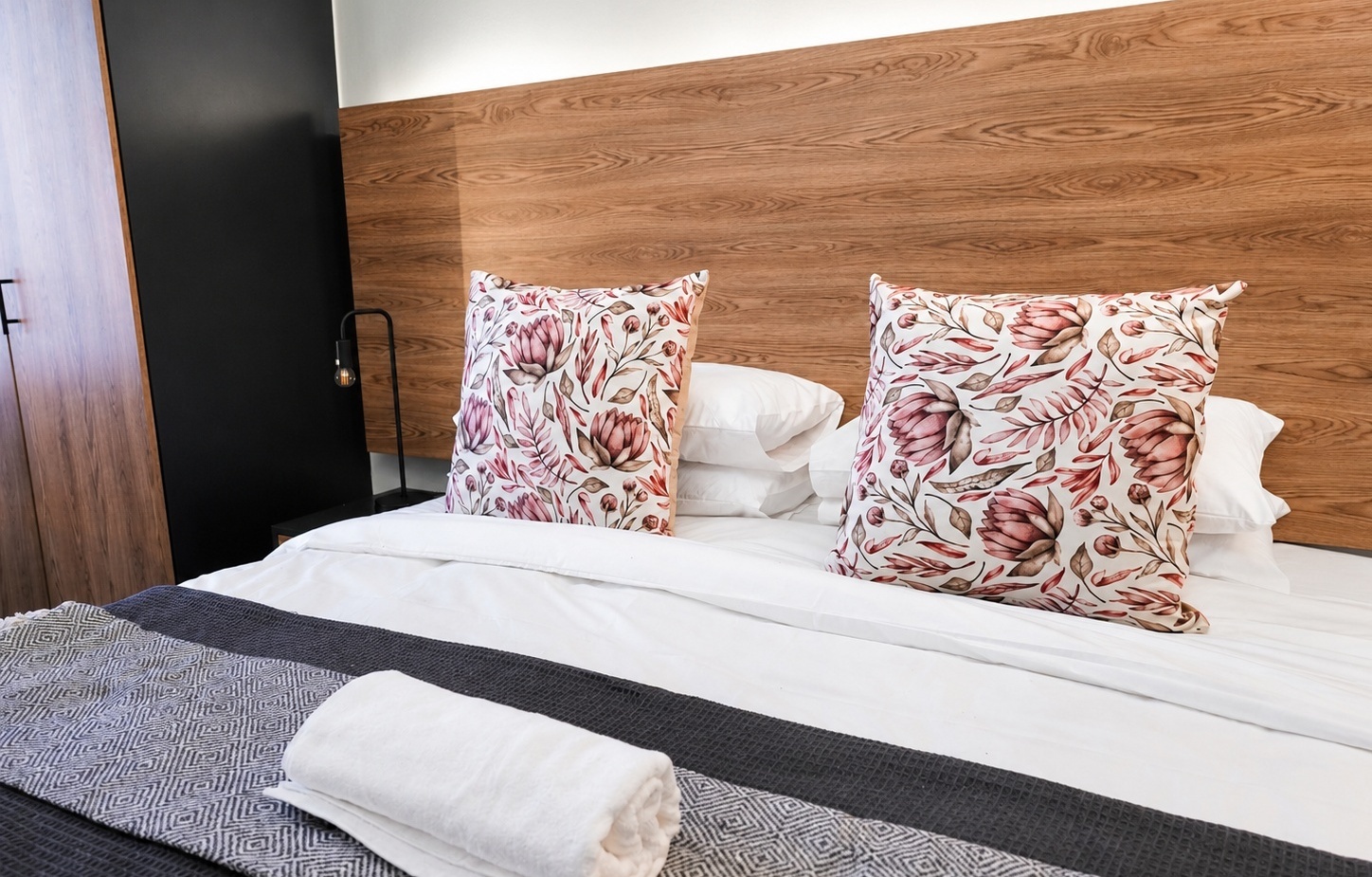Modern guestroom with wooden headboard, crisp white linens, floral decorative pillows, folded towel and textured bed runner.
