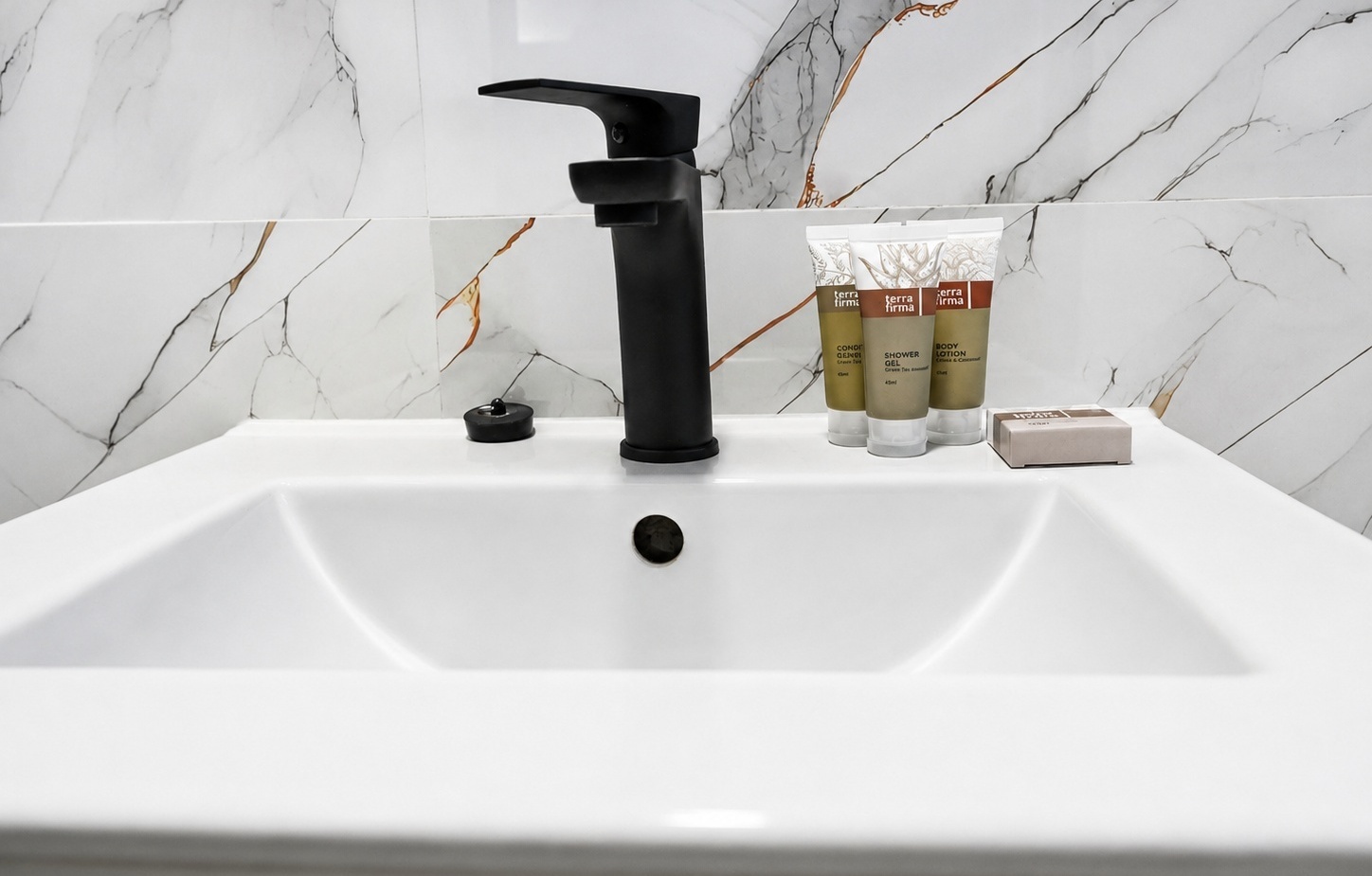 White rectangular sink with matte black faucet and marble wall, trio of hotel toiletry tubes and boxed soap on the counter.