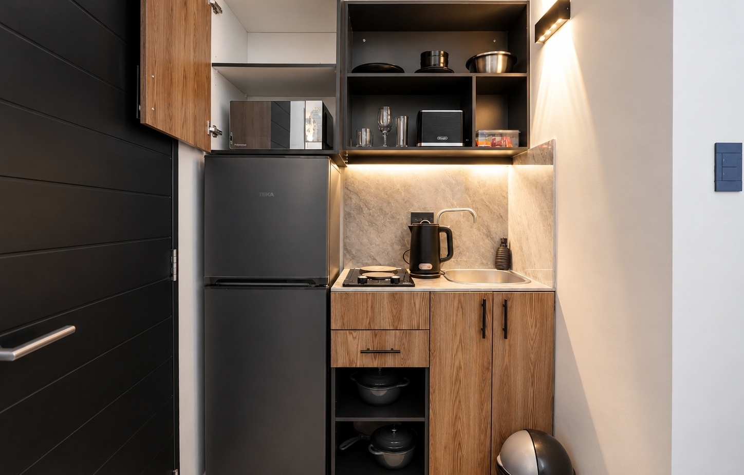 Compact kitchenette with black fridge, microwave, sink, electric kettle, hotplate, wood cabinets and under-cabinet lighting.