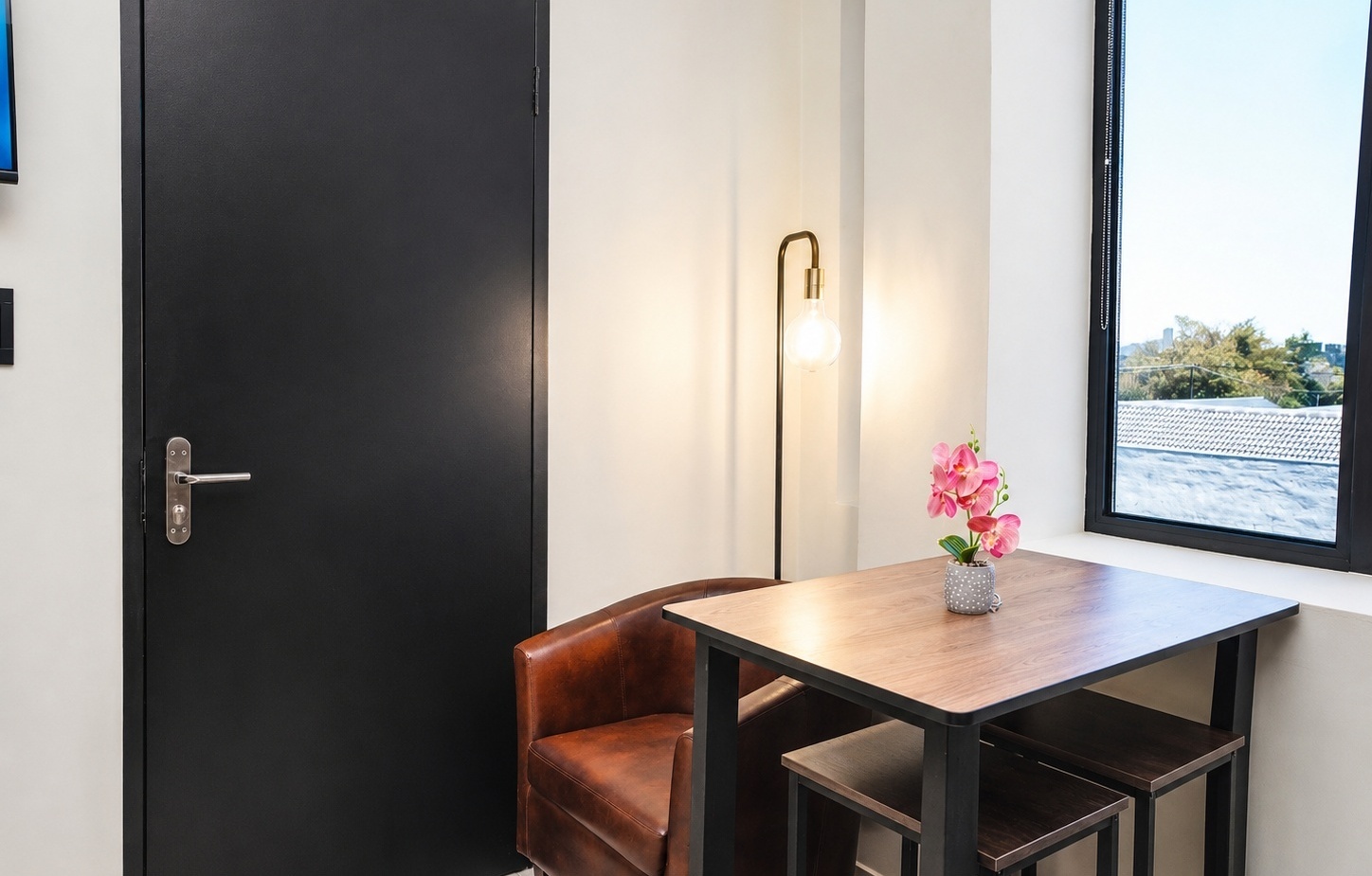 Cozy hotel nook with leather armchair, wooden high table and stools, glowing floor lamp and pink orchid by window with treetop view