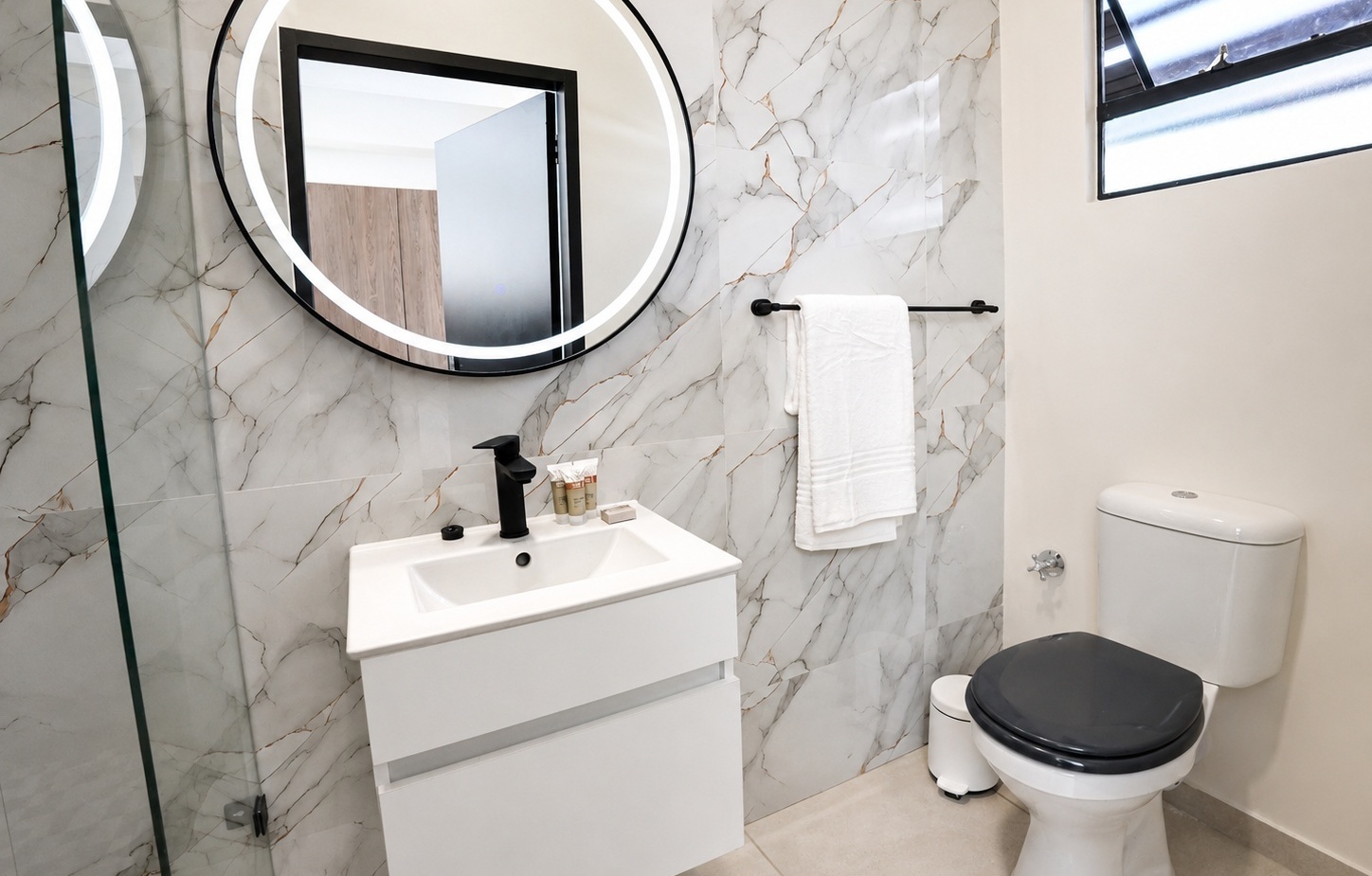 Modern bathroom with marble-look tiles, illuminated round mirror, white vanity with black faucet, toiletries and towel.