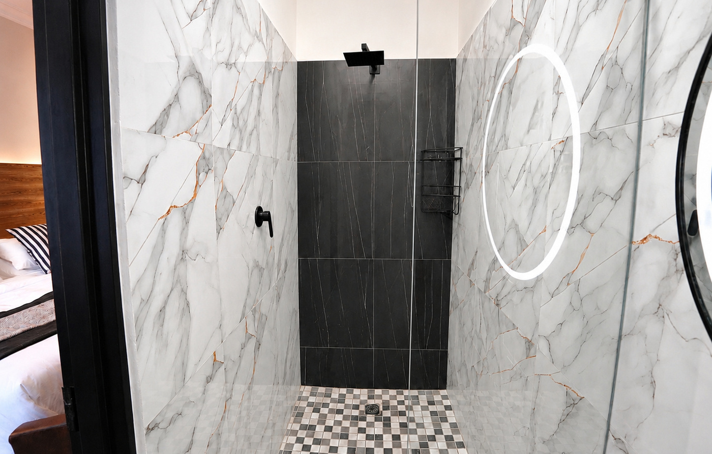 Walk-in marble shower with black rain head, mosaic floor, glass enclosure and LED-lit round mirror; view into guest bedroom.