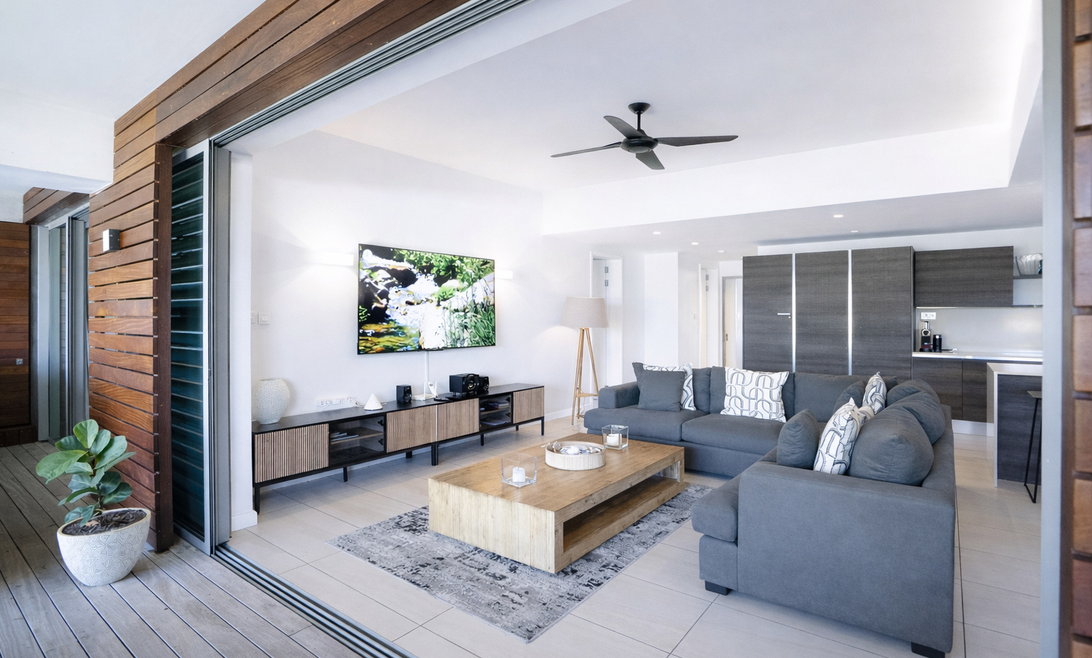 Spacious open-plan suite with sliding terrace doors, gray sectional sofa, wooden coffee table, wall-mounted TV and modern kitchenette.