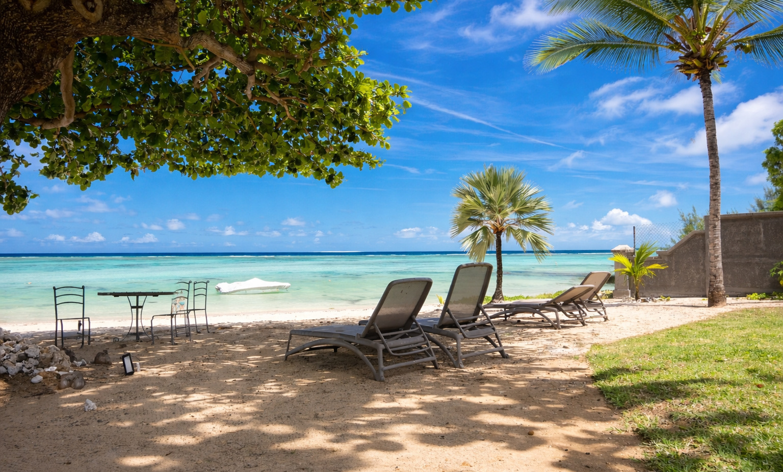 Resort's shaded beachfront with reclining loungers, small metal table and chairs on sand, palm trees and turquoise lagoon.