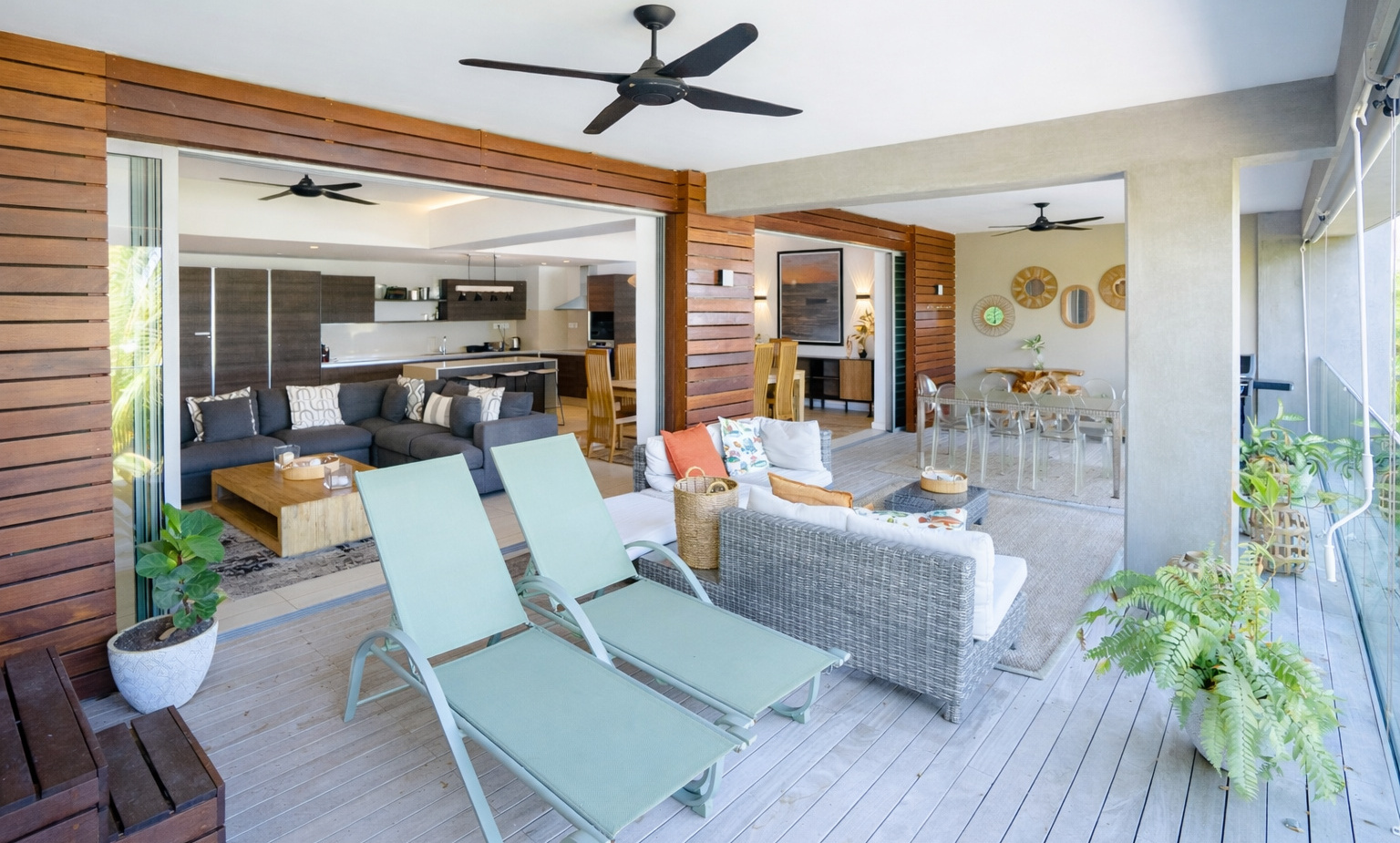 Open-plan wooden terrace with two mint sun loungers, wicker sofa, indoor lounge and dining, ceiling fans and potted plants.