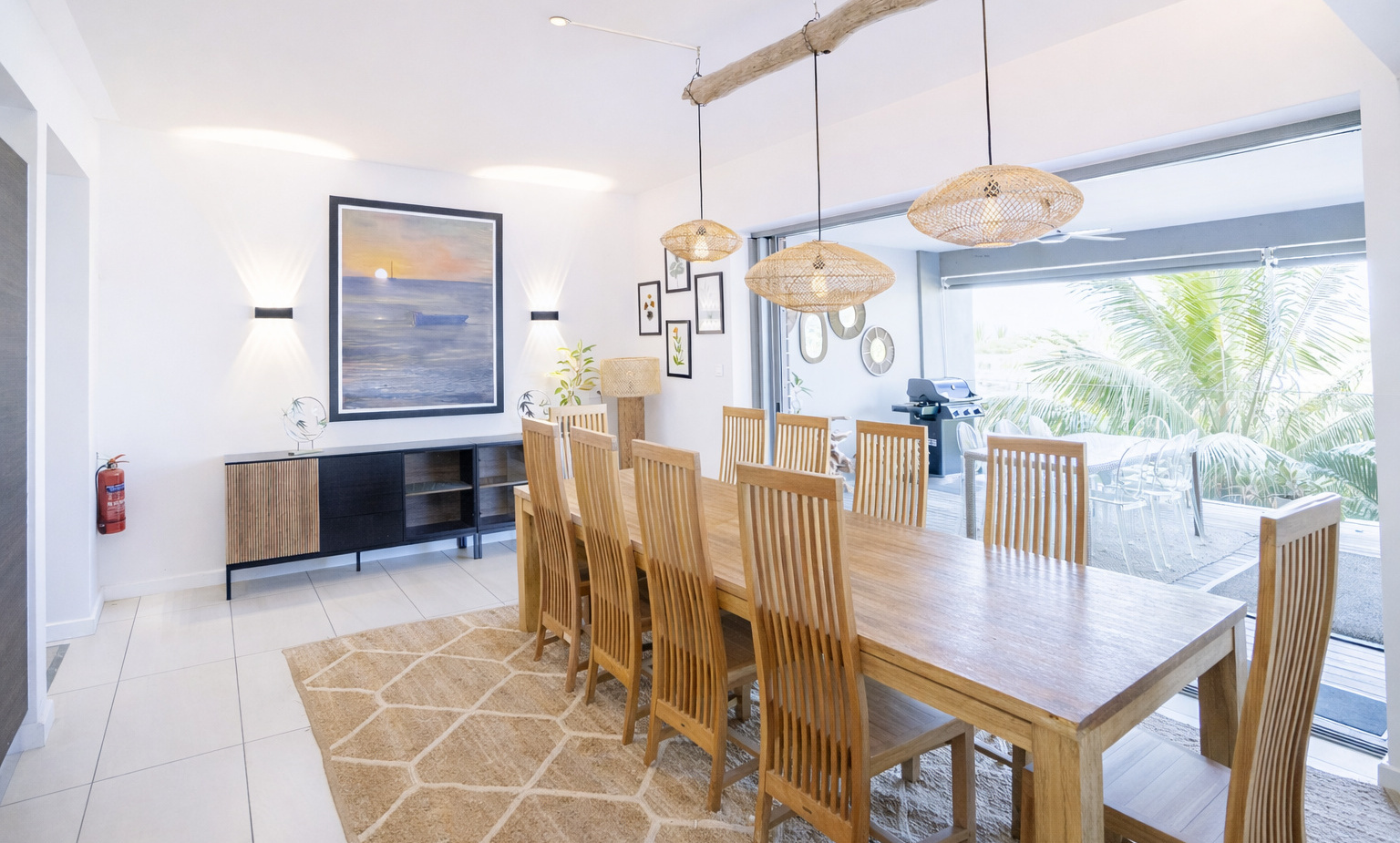 Beachfront dining room with long wooden table for ten, rattan pendant lights, ocean artwork, patio BBQ and palm view.