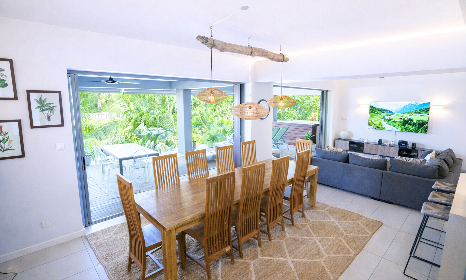 Bright open-plan dining and lounge: long wooden table for 10, rattan pendant lights on driftwood, gray sectional, sliding doors to tropical terrace