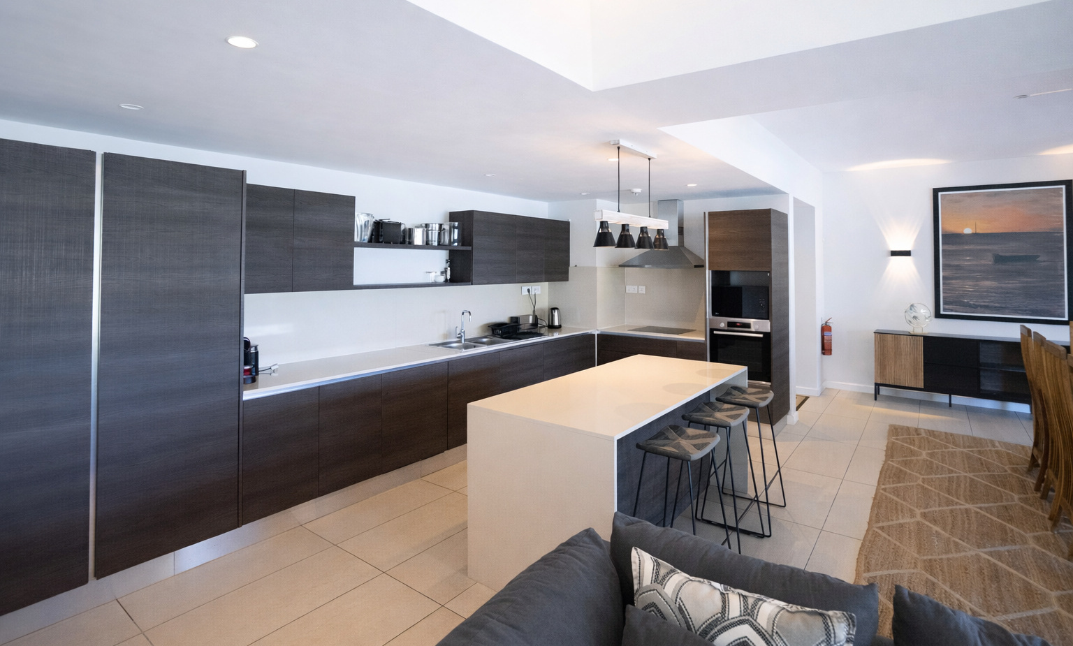 Modern open-plan hotel kitchen with dark wood cabinets, white island with bar stools, pendant lights and dining area.