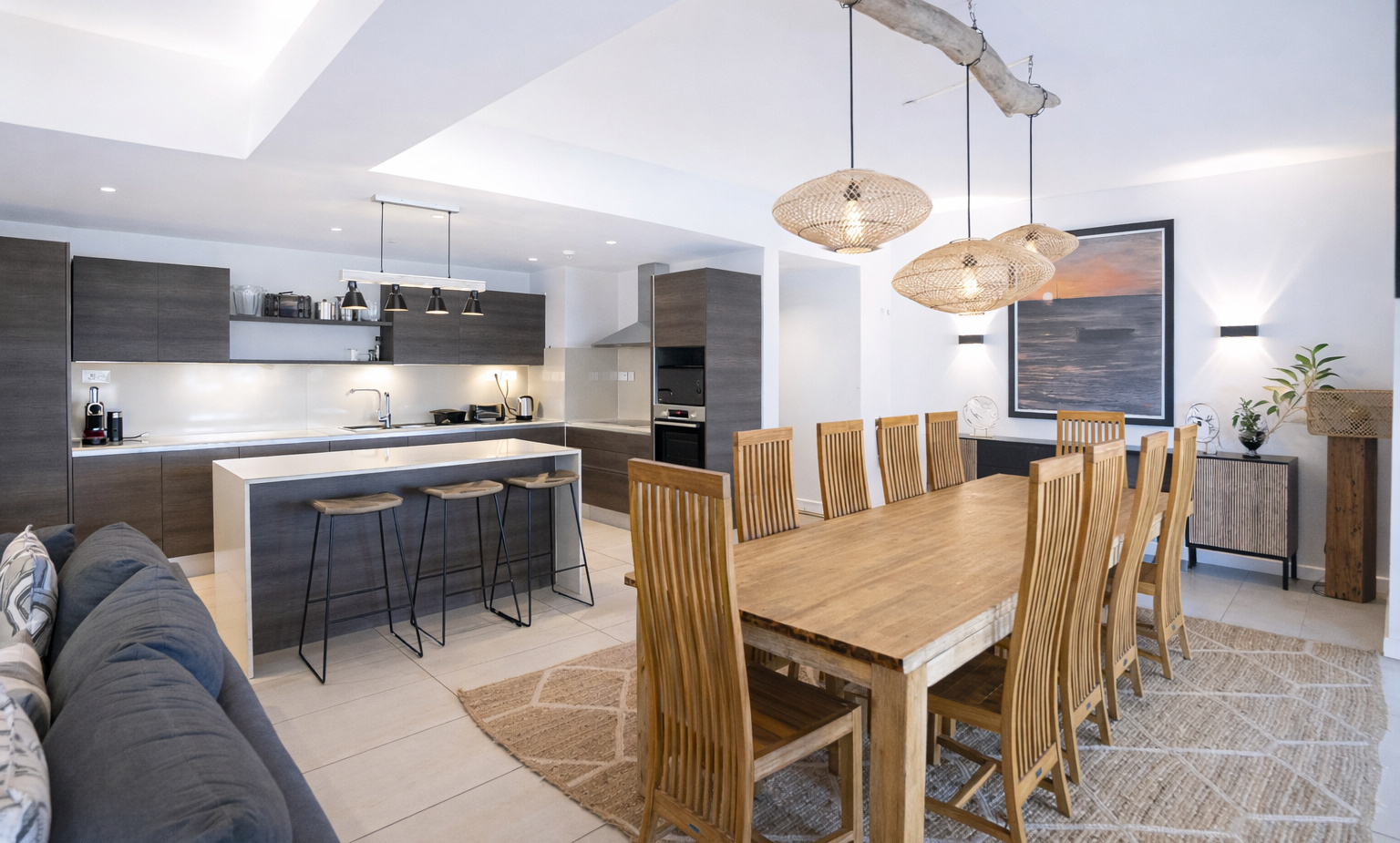 Open-plan dining and kitchen with long wooden table for 10, modern island with bar stools and woven pendant lights