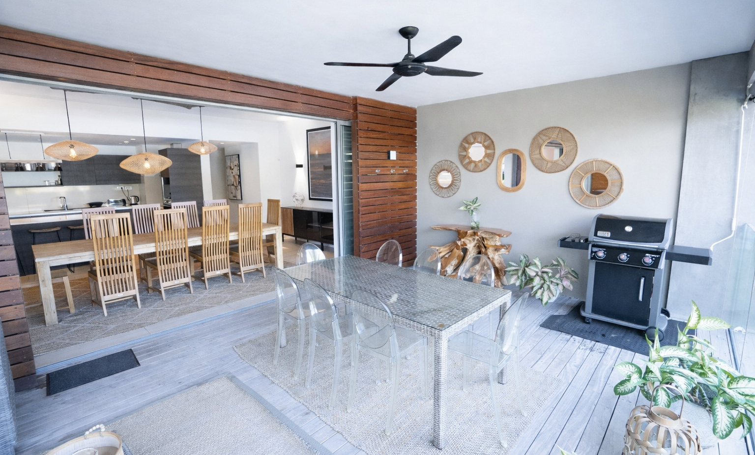 Covered terrace dining area with glass-top table and clear chairs, ceiling fan, BBQ grill, decorative rattan mirrors, open-plan indoor dining.