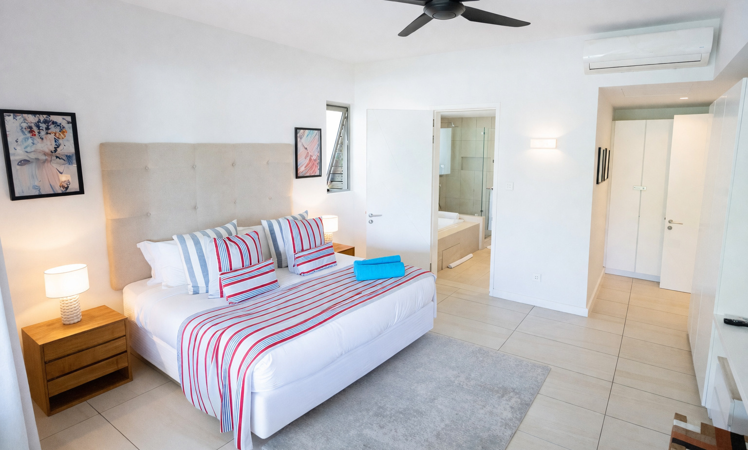 Bright modern king room with tufted headboard, red-and-blue striped bedding, bedside lamps, ceiling fan, AC and en-suite bathroom with tub.
