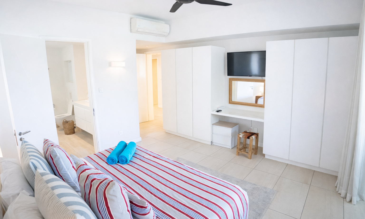 Bright modern hotel bedroom with striped bed linen, rolled blue towels, ceiling fan and air-con, built-in wardrobes, wall TV and en-suite bathroom.