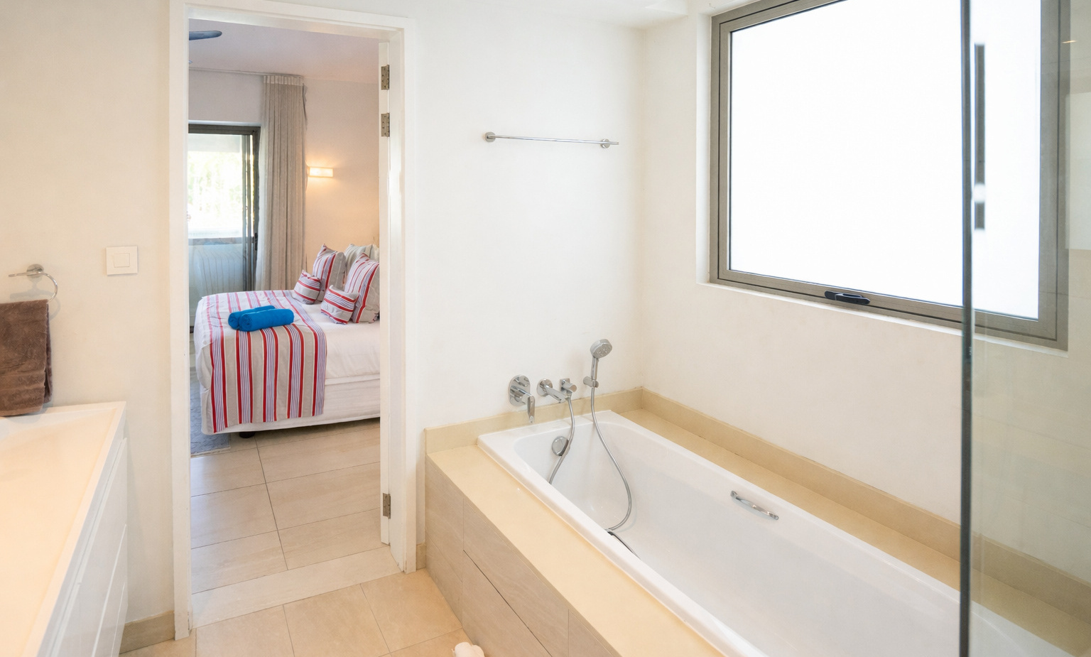 Bright modern en-suite with built-in bathtub and handheld shower, frosted window, and view into bedroom with striped bedding.