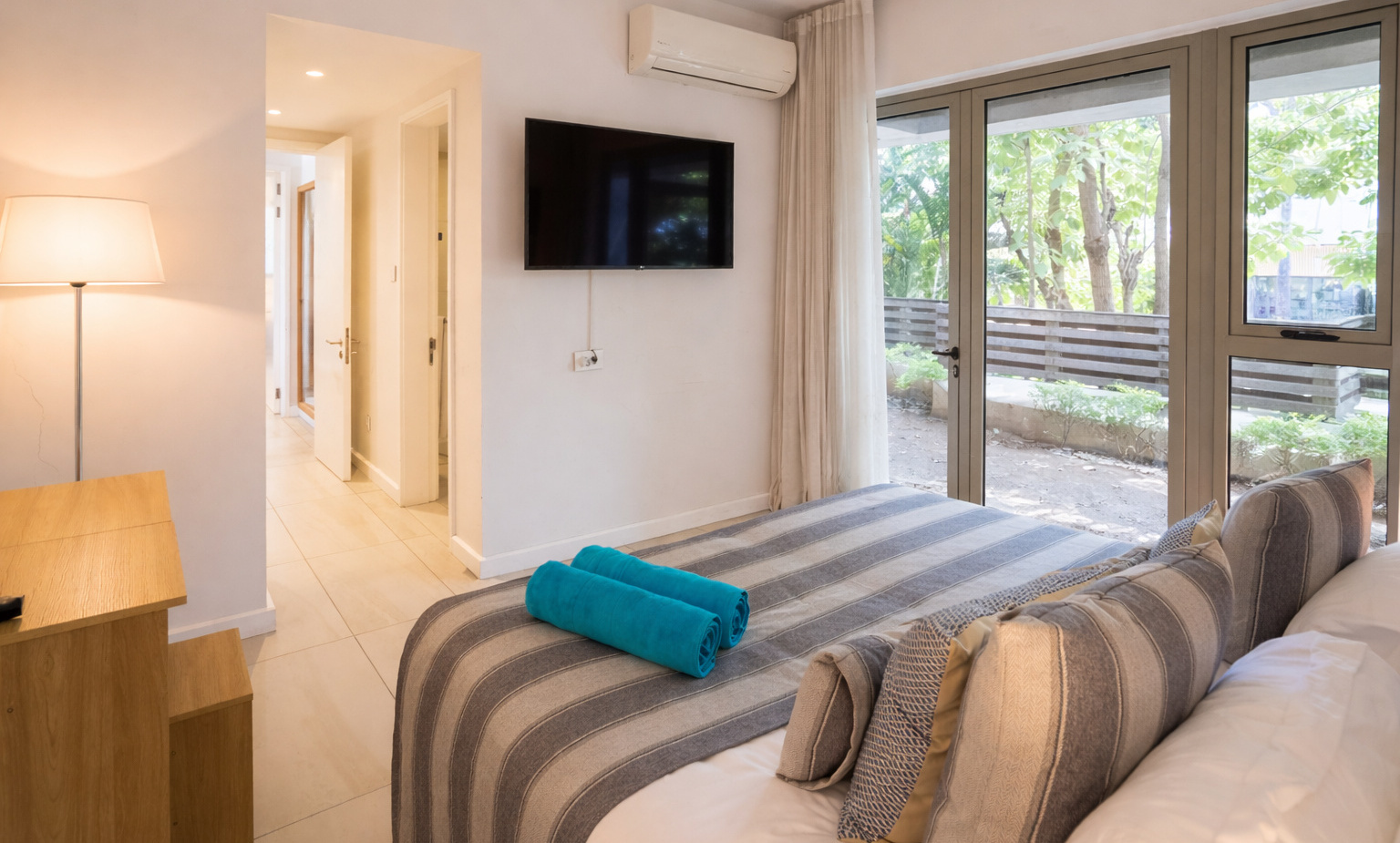 Ground-floor room with striped king bed, turquoise towels, decorative pillows, wall TV, desk, AC, glass doors to leafy patio.