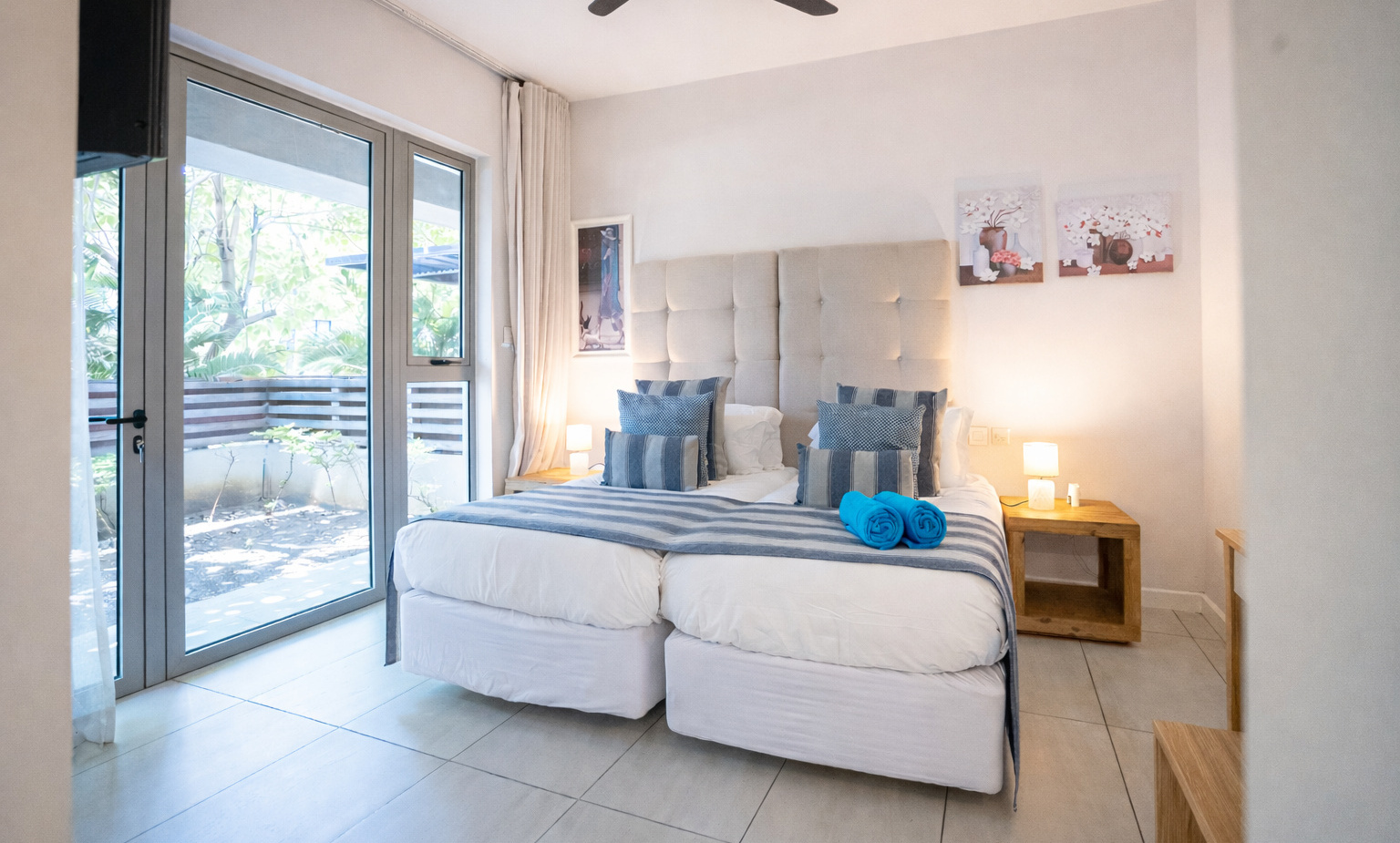 Bright ground-floor twin room with tufted headboard, glass patio doors to leafy terrace, striped blue linens, bedside lamps and rolled towels.