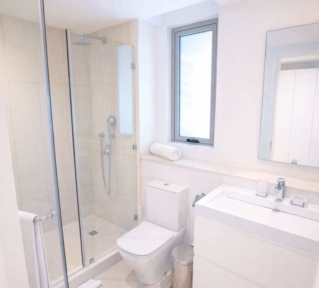 Bright modern hotel bathroom with glass walk-in shower (rain and handheld showerheads), white vanity, toilet, rolled towel on window ledge.